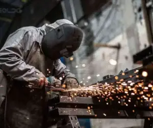 Caracterização de Materiais 6 Trabalhador realizando análise de superfície metálica com faíscas visíveis.