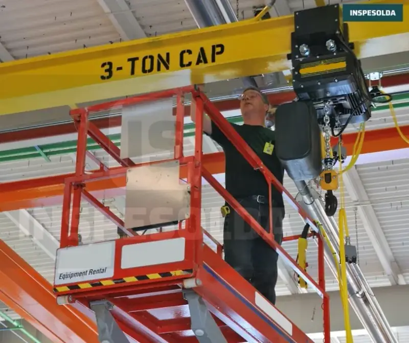Técnico em plataforma elevatória examina a talha elétrica presa à viga amarela de uma ponte rolante de 3 toneladas em galpão industrial.