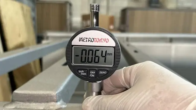Mão com luva segurando relógio comparador digital diante de uma estrutura metálica.