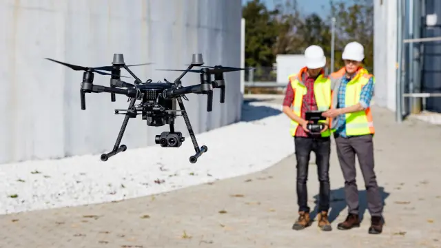 Drone em voo diante de tanques industriais, com duas pessoas operando o controle remoto.